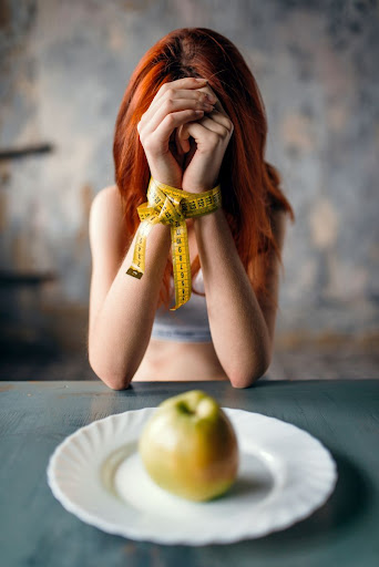 žena si zakrýva tvár rukami, zviazanými meradlom, pred sebou má tanier s jablkom